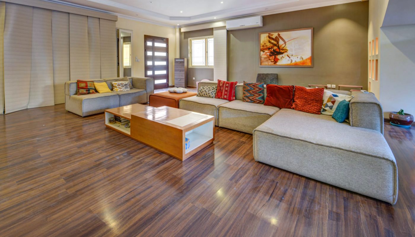 Spacious living room with wooden floors, light grey sectional sofas, colorful pillows, a wooden coffee table, large windows, and a painting on the wall.