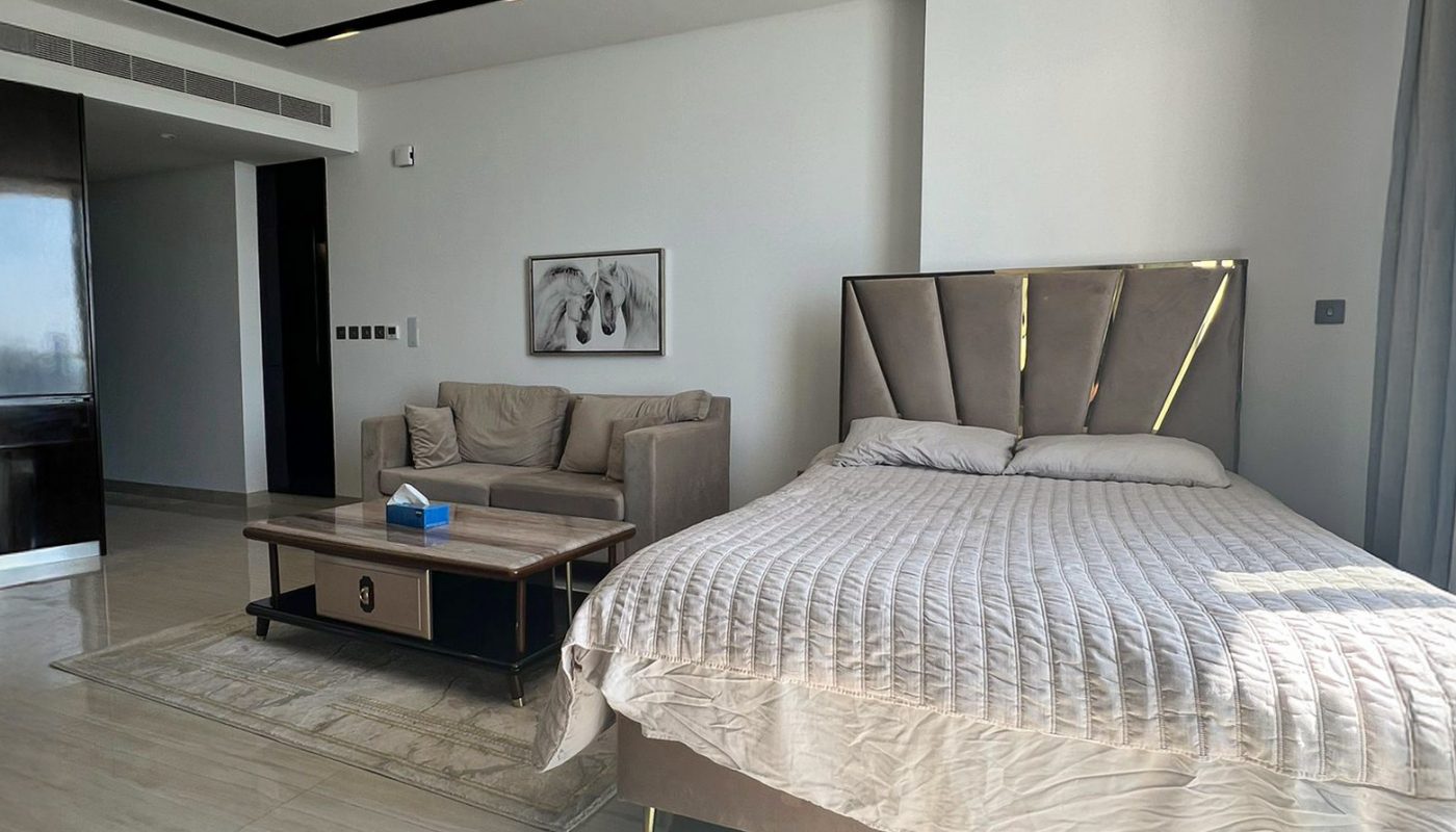 A modern bedroom with a large bed, a small sofa, a coffee table, a wall painting, and light-colored tile flooring. Sunlight is streaming in through the window.