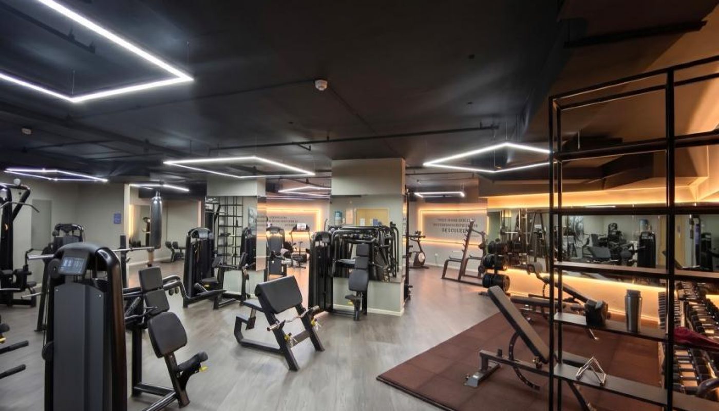 Spacious modern gym with various exercise machines, free weights, and mirrors, featuring wooden flooring and geometric ceiling lights.