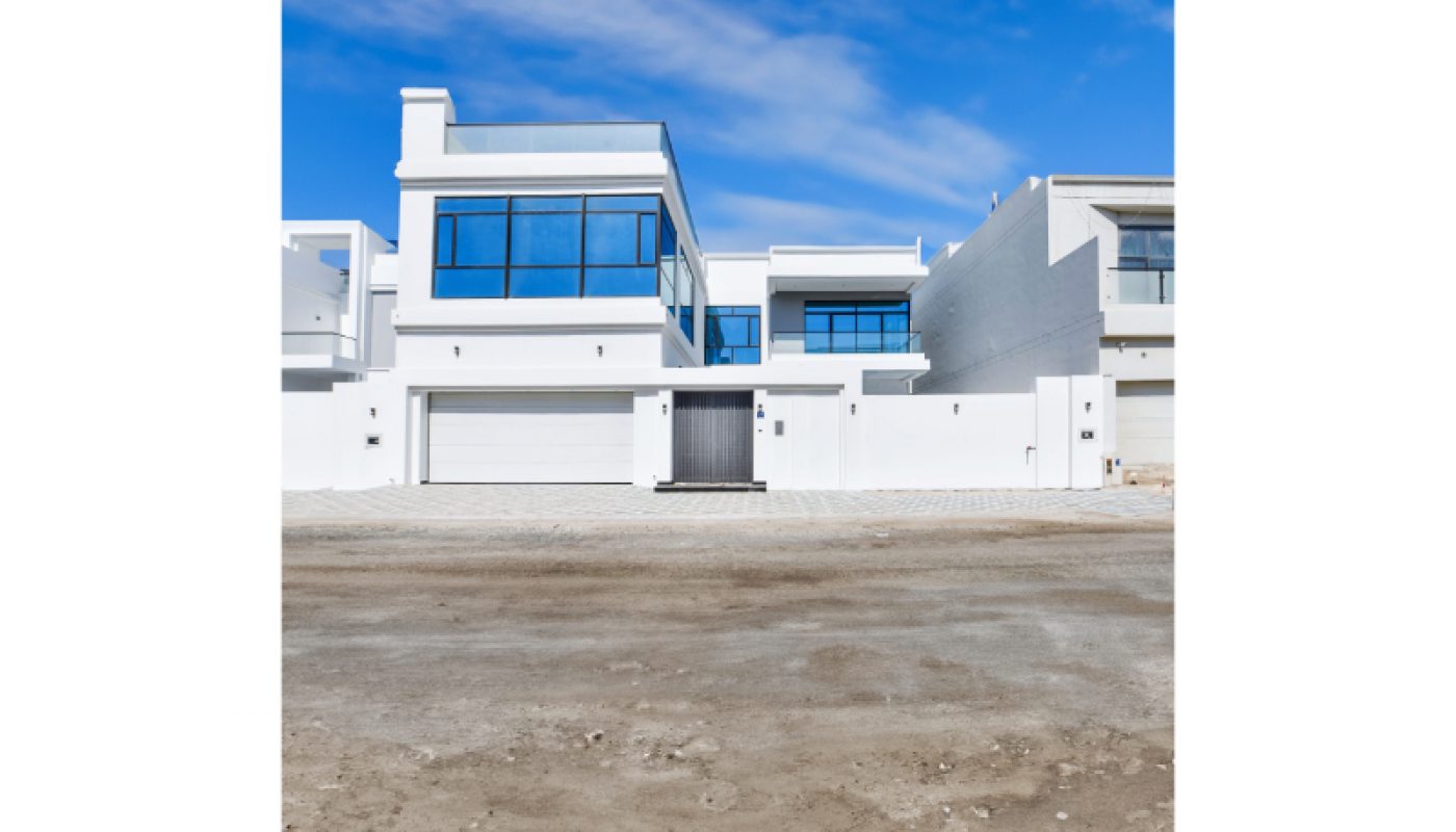 Modern white two-story house with large blue-tinted windows, a double garage, and a paved driveway, viewed from across an unpaved road under a blue sky.