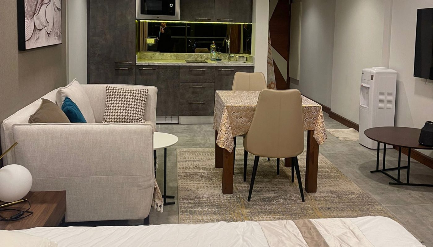 A modern studio apartment features a bed, sofa, dining table, kitchenette, TV, and wall art in a well-lit, open-plan layout.