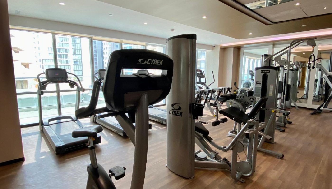 Modern gym room with large windows, containing treadmills, stationary bikes, and various exercise machines on a wooden floor.