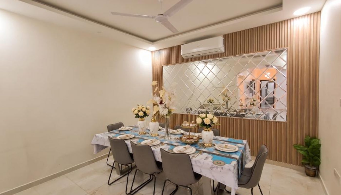A modern dining room with a table set for eight, decorated with floral centerpieces, grey chairs, and a large mirrored wall with wooden paneling.