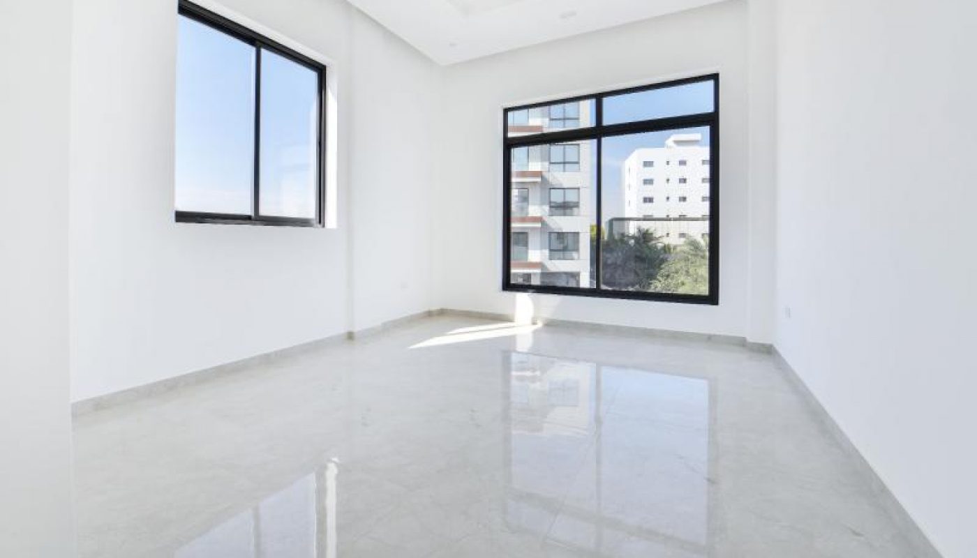 Empty, unfurnished room with white walls, light-colored tiled floor, and two large windows showing a view of nearby buildings and trees.