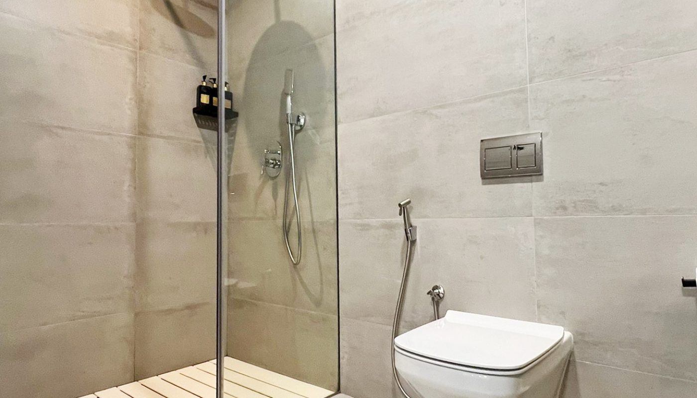 Modern bathroom with a walk-in shower, wall-mounted toilet, handheld bidet, and a light-colored waste basket on a tiled floor and walls.