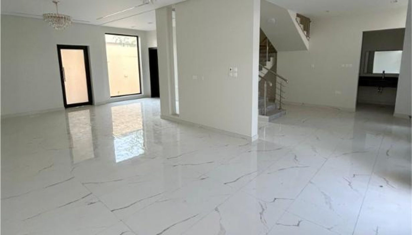 Spacious, empty room with white marble floors, modern chandeliers, staircase, and neutral walls; large windows and open doorways visible.