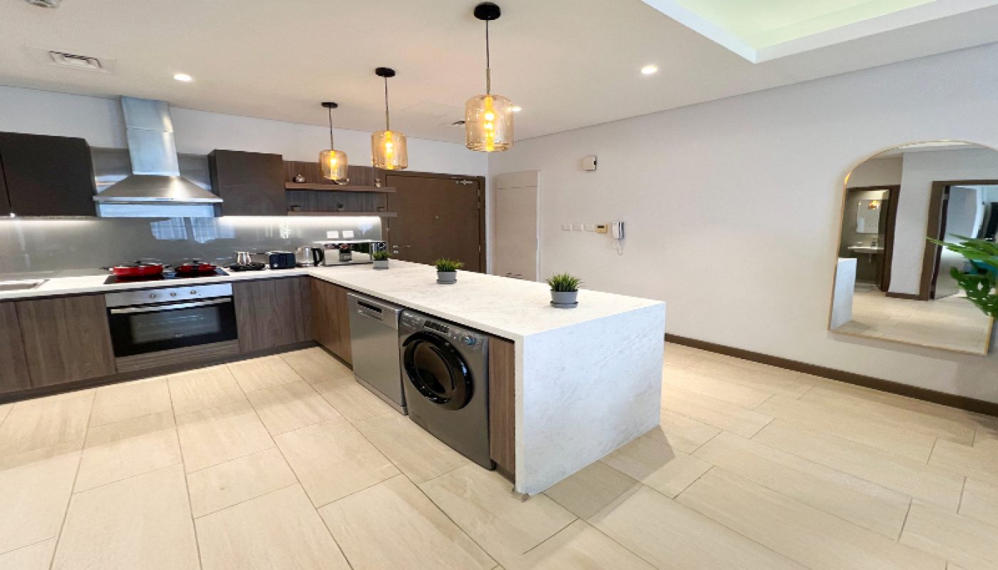 Modern kitchen with an island featuring a built-in washing machine, pendant lights, dark cabinets, light countertops, and a wall mirror reflecting a hallway.