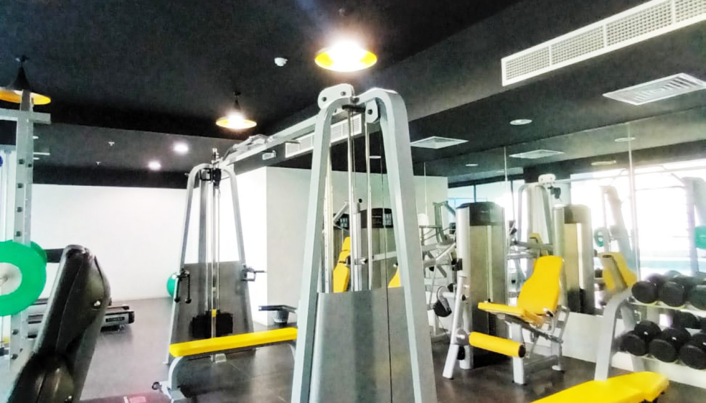 A gym with exercise machines, yellow benches, weight racks, and mirrored walls under ceiling lights.