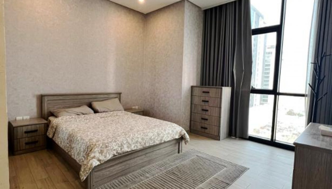 A modern bedroom with a double bed, two dressers, a large window with curtains, and light wood flooring.