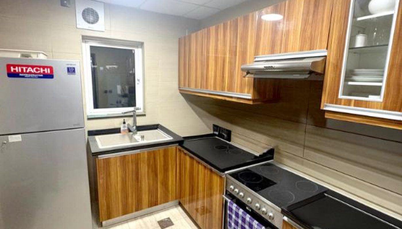Modern kitchen with wood-finish cabinets, stainless steel appliances, electric stove, fridge, sink, and light-colored tiled floor. A window provides natural light above the sink.