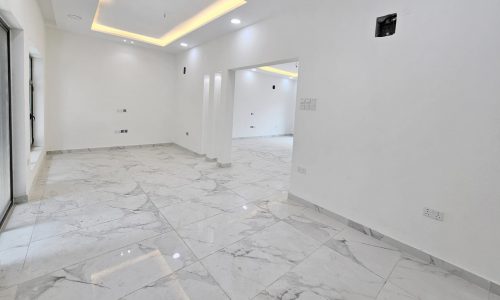 A spacious, empty room in a luxurious villa with white walls, marble flooring, a recessed ceiling with built-in lighting, and large windows on the left side. Perfect for those seeking elegance and comfort. Villa for Sale in the prestigious Barbar neighborhood.