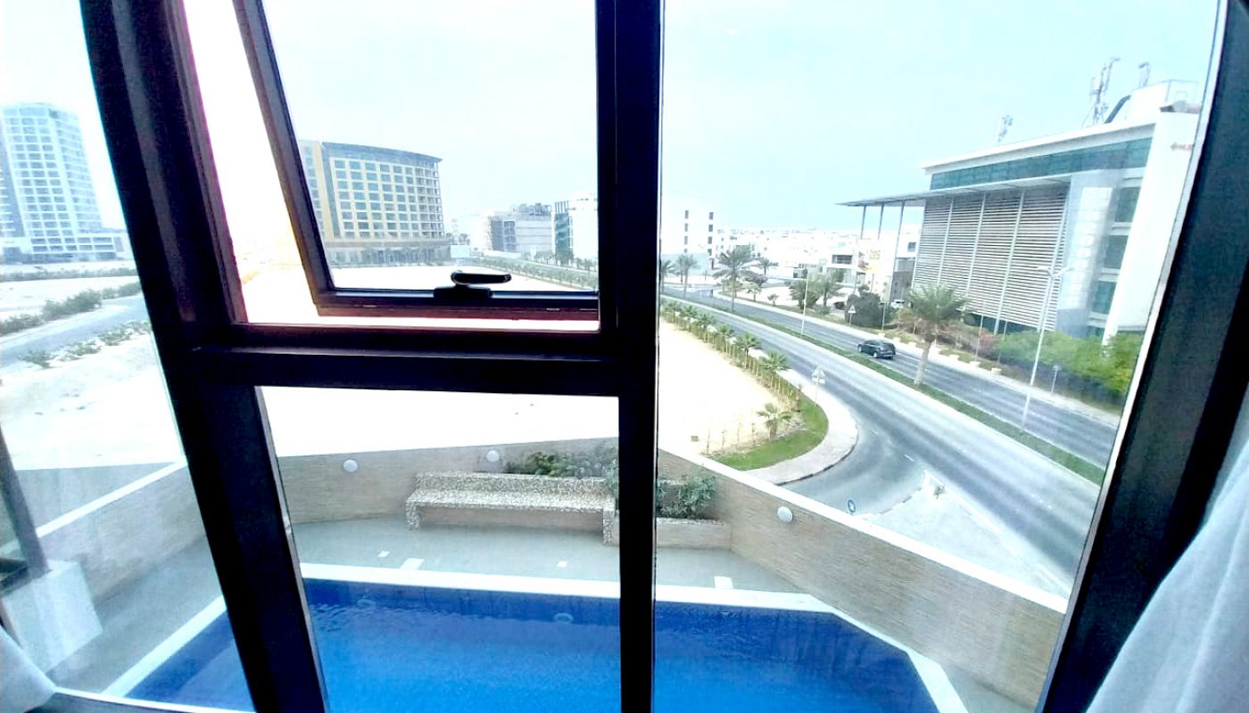 View through a large window showing a blue swimming pool below, a curved road, and modern buildings in the background under an overcast sky.