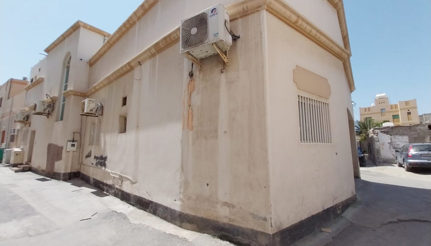 A beige two-story building with barred windows and air conditioning units, located on a quiet street under a clear sky.