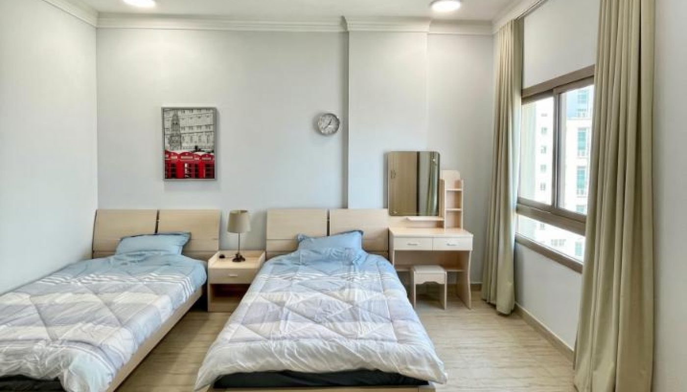 A bright, tidy room with two single beds, a small desk with a mirror, a chair, a lamp, a clock on the wall, and a large window with beige curtains.