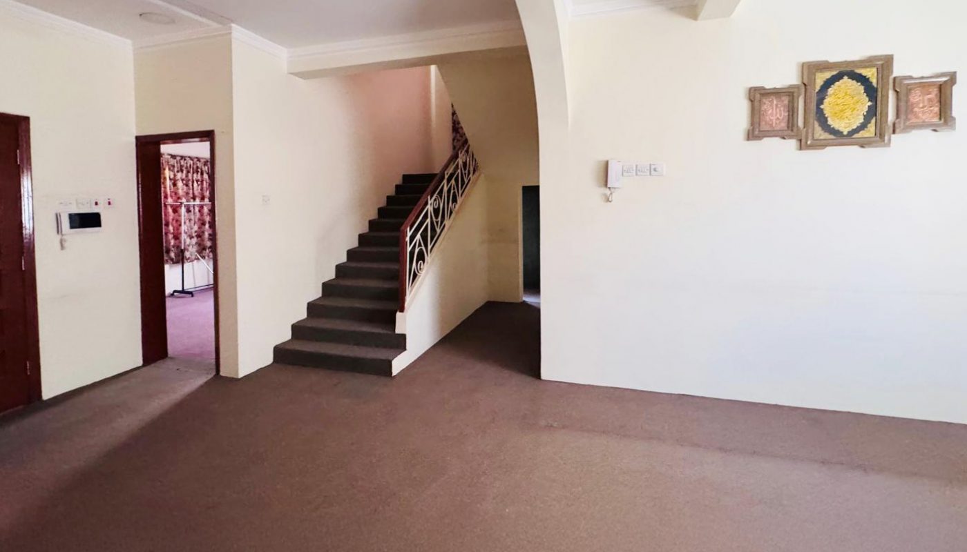 A spacious, empty room with beige walls, a brown carpet, a ceiling fan, a staircase in the center, and wall art on the right. Several doorways lead to other rooms.