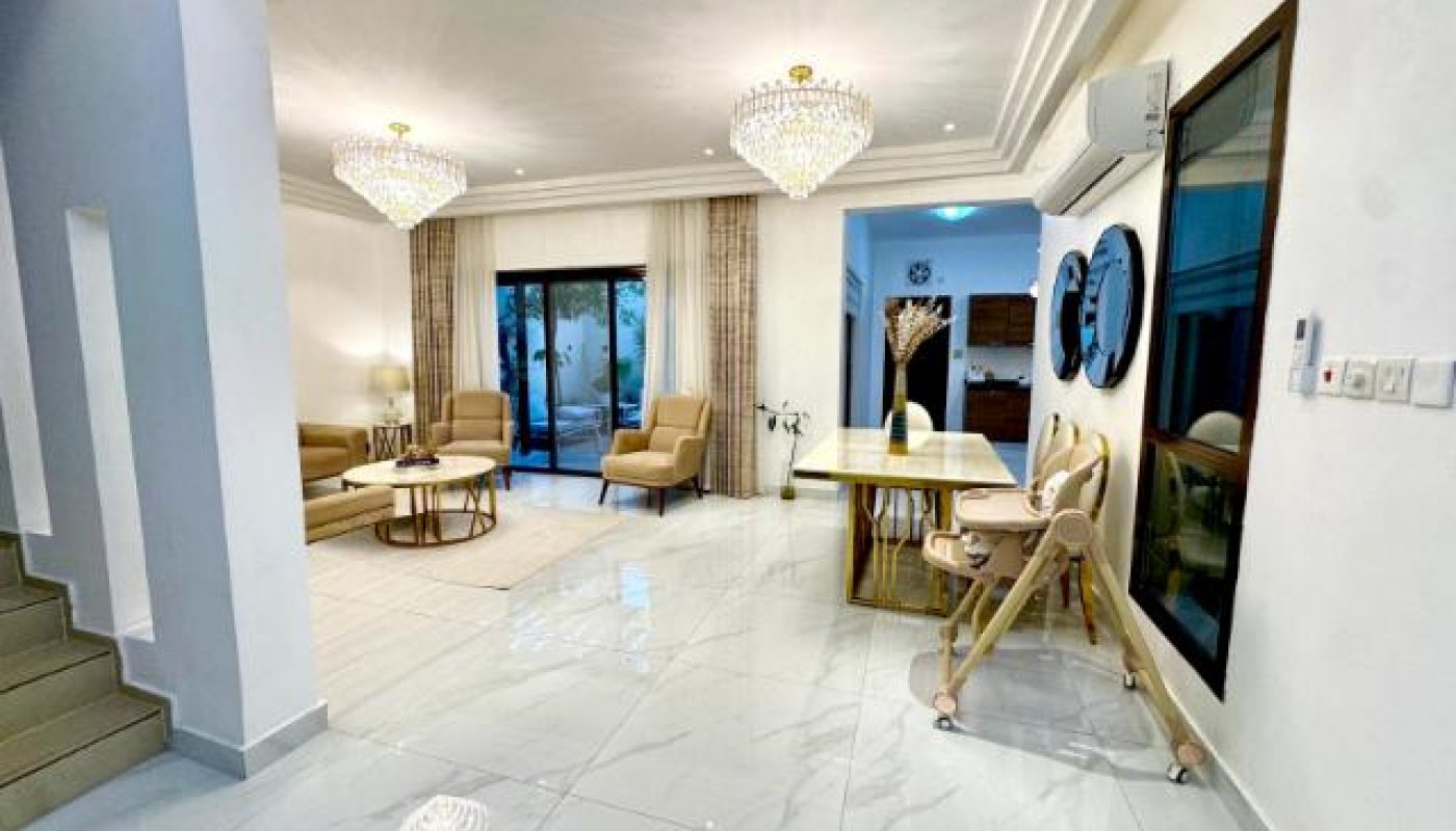Modern, open-plan living and dining room with marble floor, neutral furniture, chandeliers, and large windows. A high chair and mirrors are near the dining table. Stairs on the left.
