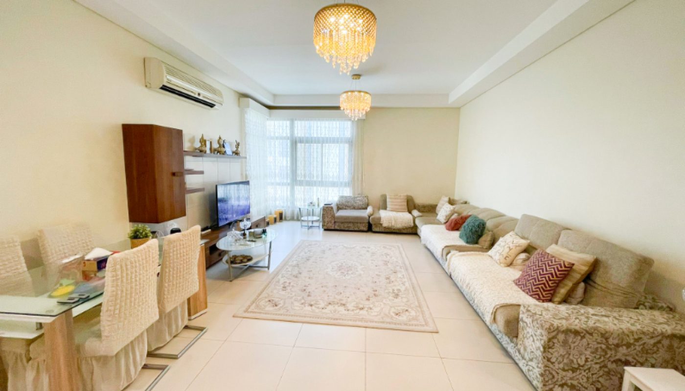 Spacious living room with neutral tones, a large rug, sectional sofas, dining table, TV, chandeliers, and a window with sheer curtains.