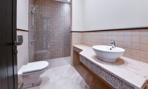 A modern bathroom in an Al Areen 3BR villa for sale features a walk-in shower, a white toilet, a vessel sink on a spacious countertop, and beige tiled walls.