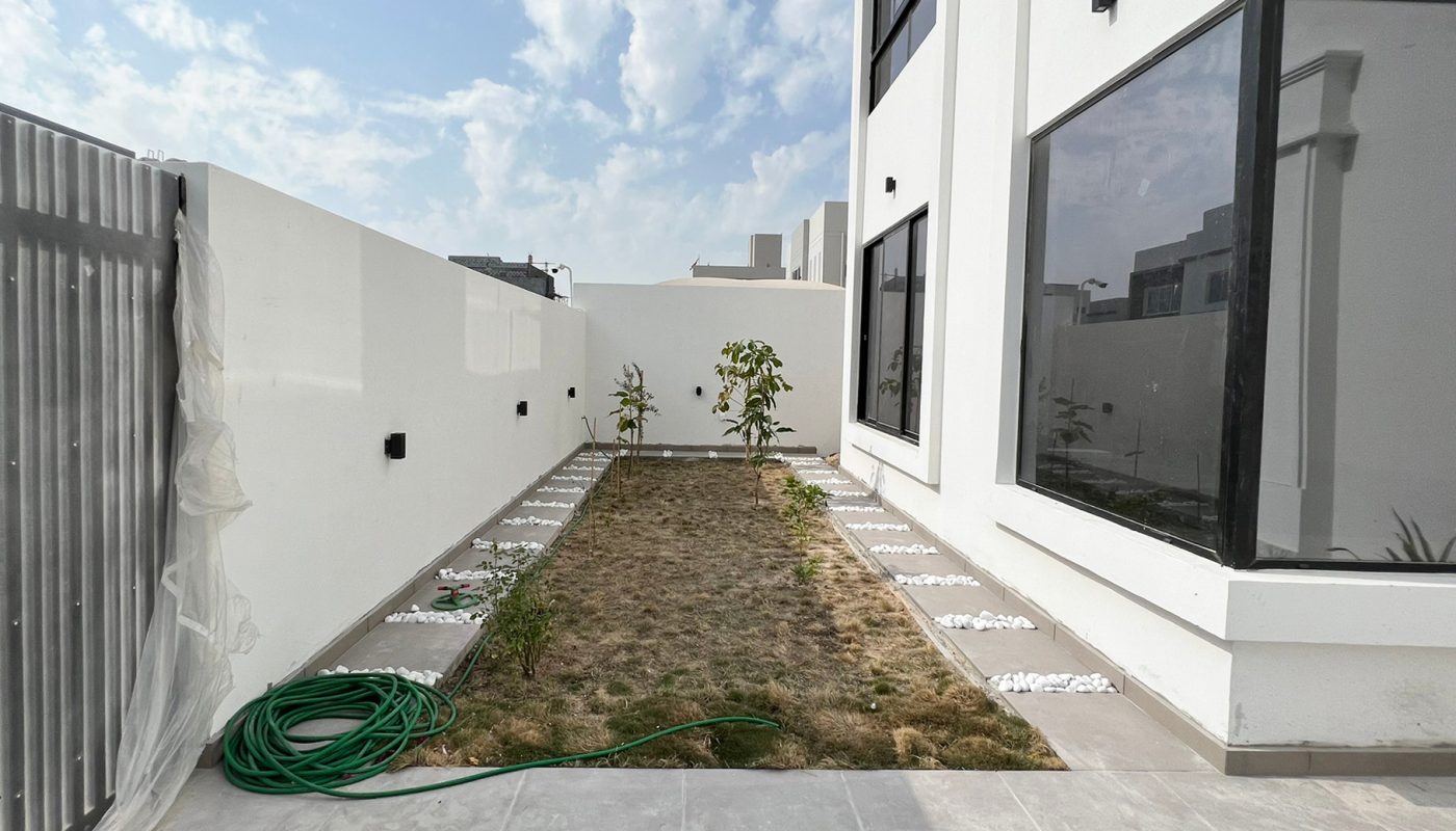 A modern white building with large windows and a small yard featuring dry grass, a green garden hose, and young plants along the perimeter, under a partly cloudy sky.