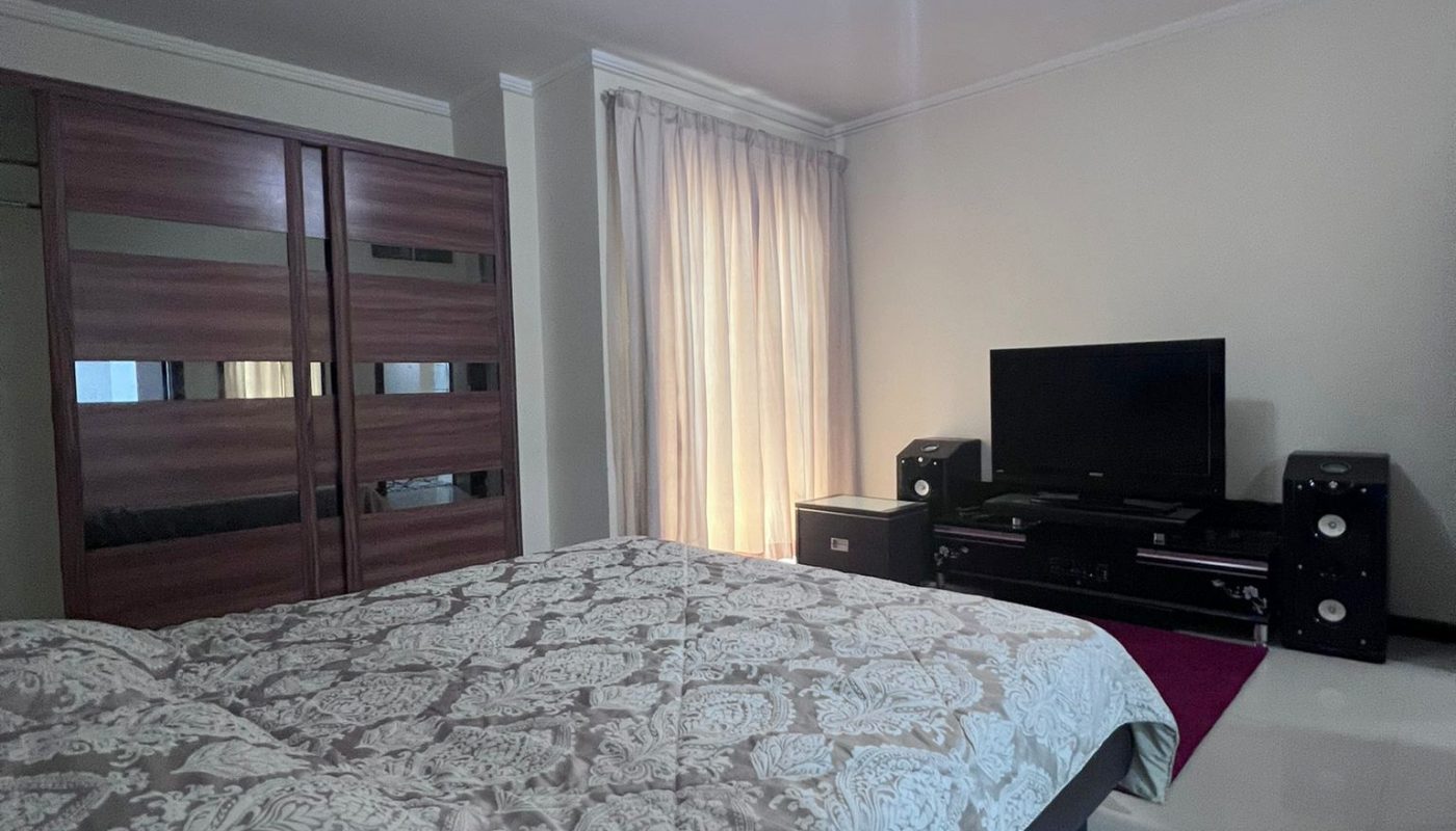Bedroom with a patterned bedspread, large wardrobe, curtained window, and a TV with speakers on a low cabinet against a white wall.