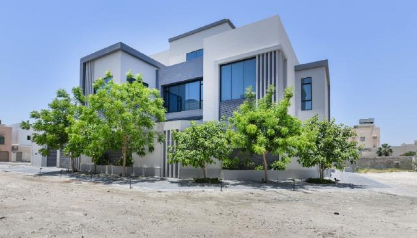 Modern white two-story building with large windows, surrounded by several green trees, set on a mostly bare, sandy lot under a clear blue sky.