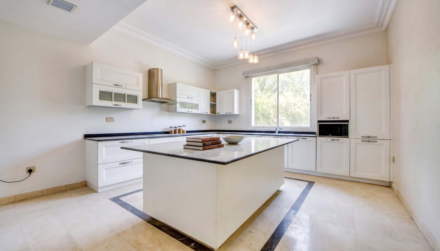 Modern kitchen with white cabinets, black countertops, a central island, overhead lights, and a large window offering natural light. Marble floor and minimalist décor are visible.