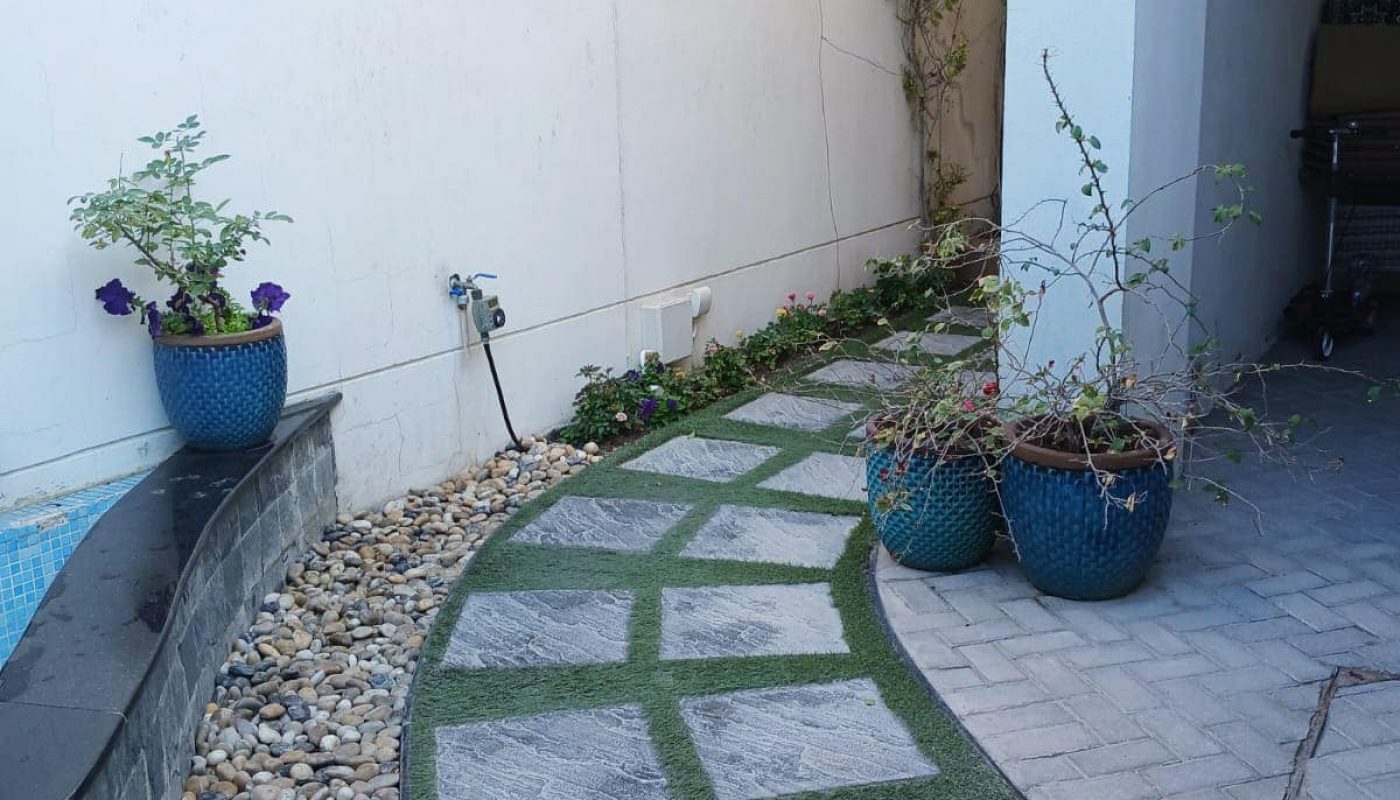 A landscaped garden path with stone tiles and artificial grass, lined by potted plants and a pebble border beside a white wall.