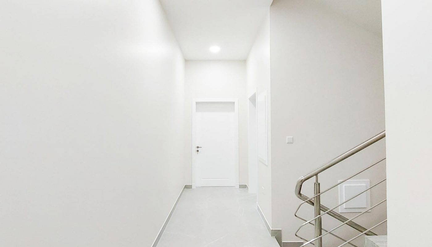 A bright, minimalist hallway with white walls, a closed white door at the end, and a metal handrail beside a staircase on the right.
