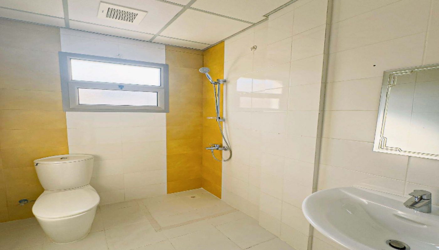 A bathroom with a toilet, wall-mounted shower, sink with a faucet, mirror, and a window. The walls are tiled in white and yellow.