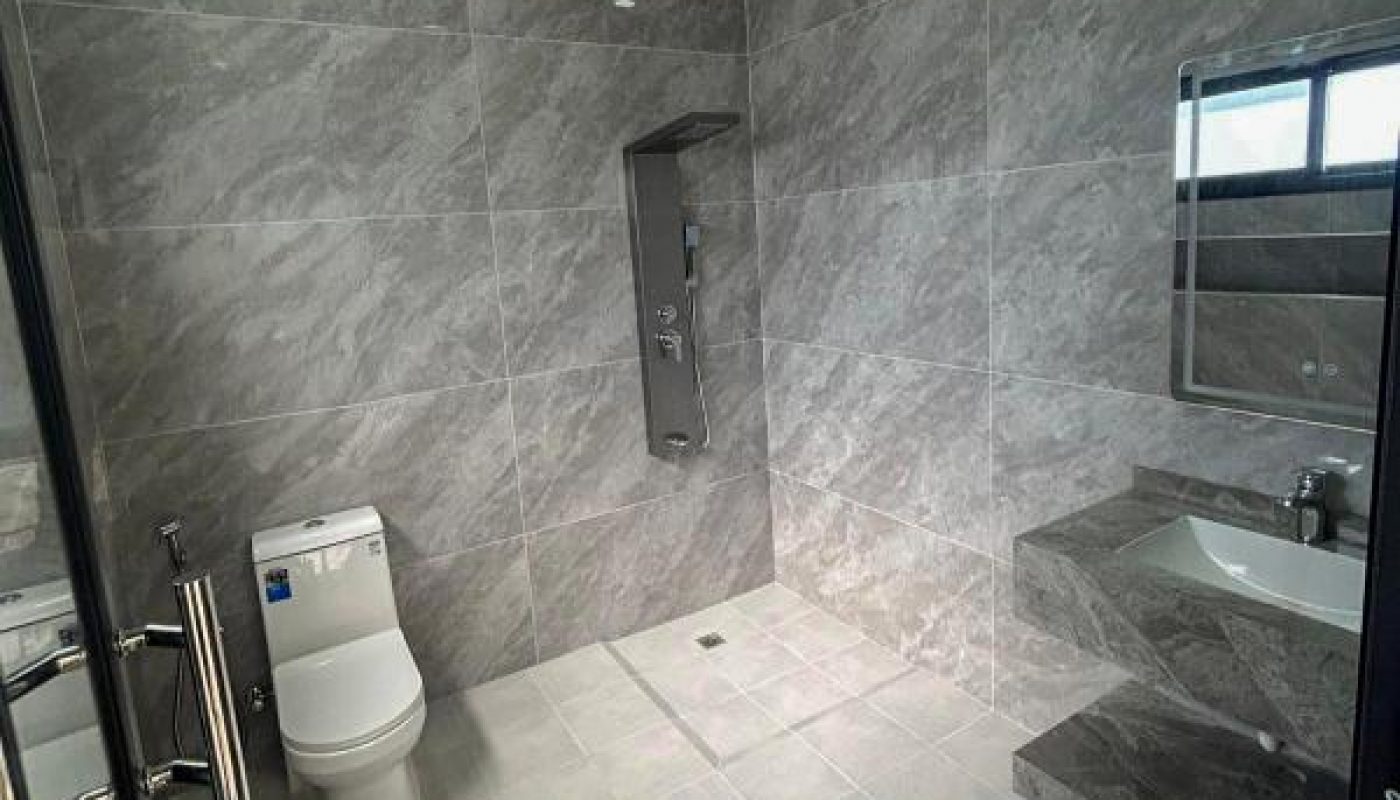 A modern, spacious bathroom with gray tiled walls and floor, featuring a toilet, a wall-mounted sink with a mirror, and a shower area.
