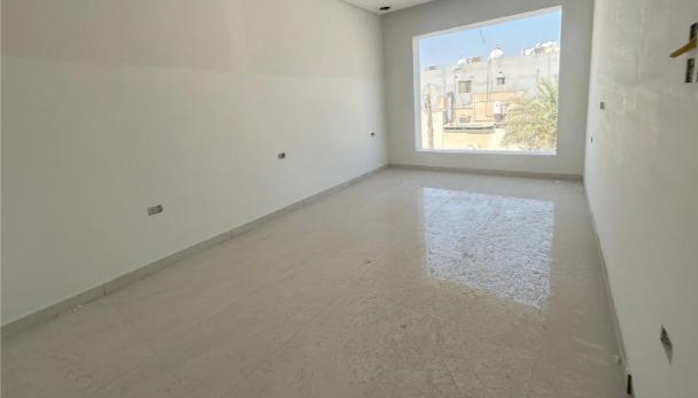 Empty, unfurnished room with white walls, tiled floor, and a large window showing buildings outside. Ceiling has a grid pattern with some visible fixtures and wiring.