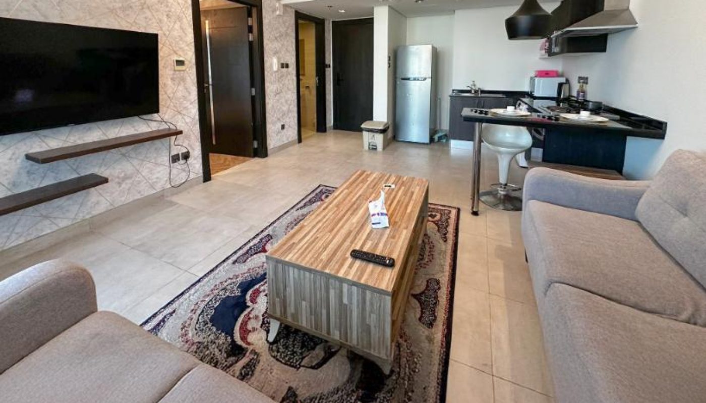 Modern living room with two gray sofas, a wooden coffee table, wall-mounted TV, patterned rug, and open kitchen area with barstools and a refrigerator in the background.