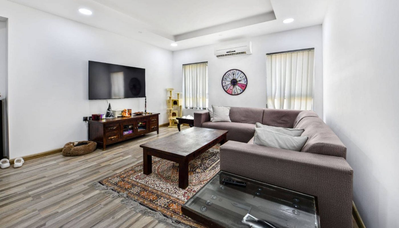 A modern living room with a large gray sectional sofa, flat-screen TV, and a wooden coffee table on a patterned rug. Air conditioner and artwork on the wall. Wood-style flooring.