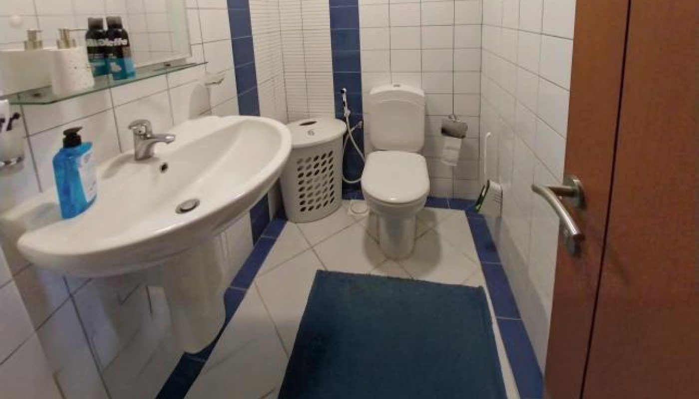 A bathroom with white tiled walls, a sink with toiletries, a blue mat, a toilet, a laundry basket, and a mirror above the sink.