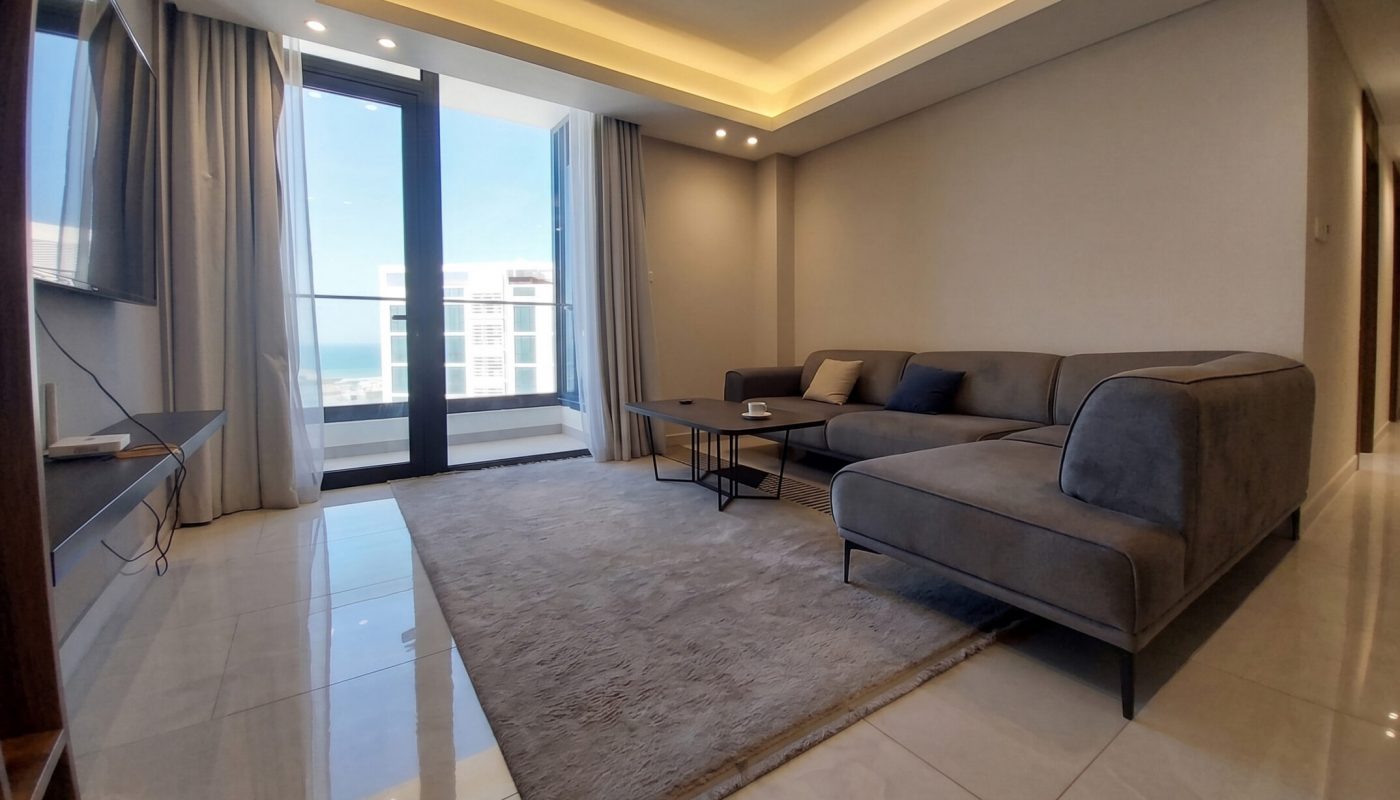 Modern living room with a grey sectional sofa, rug, television, and large windows letting in natural light; neutral tones and minimalistic decor.