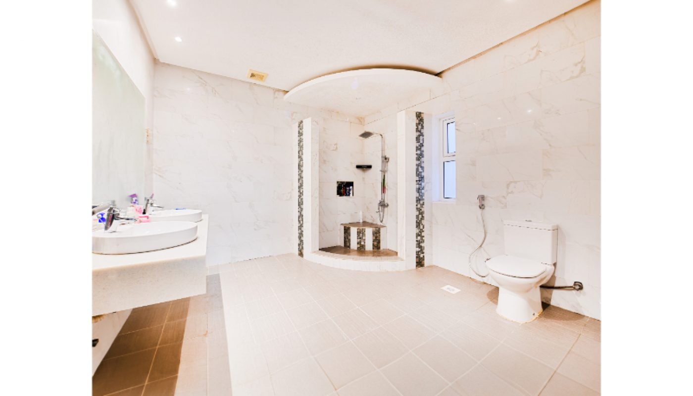 Spacious bathroom with tiled floors and walls, a walk-in shower, toilet, and a double sink vanity with toiletries on the counter.