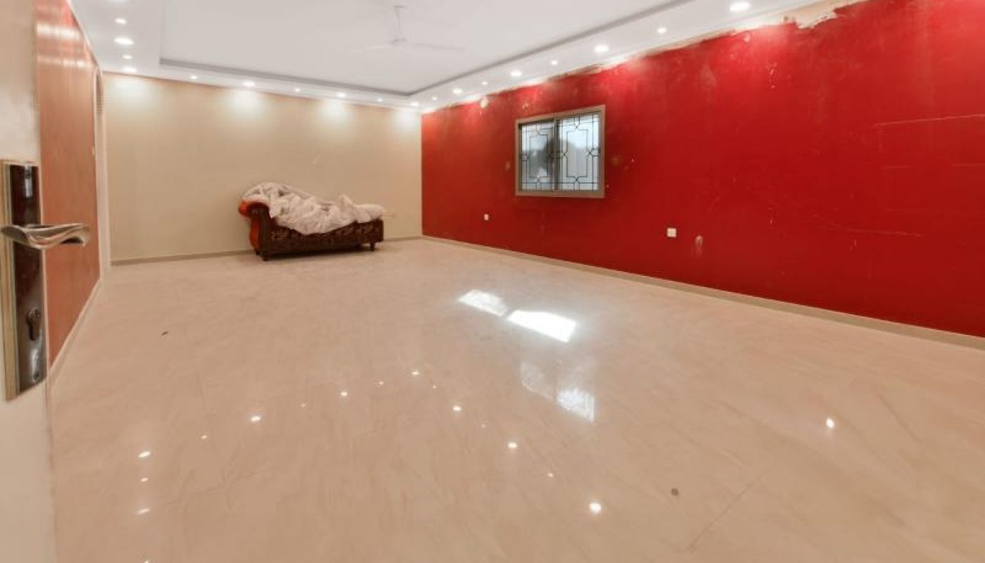 Spacious, empty room with beige tiled floor, red and white walls, ceiling lights, a window, and a single couch covered with a sheet.