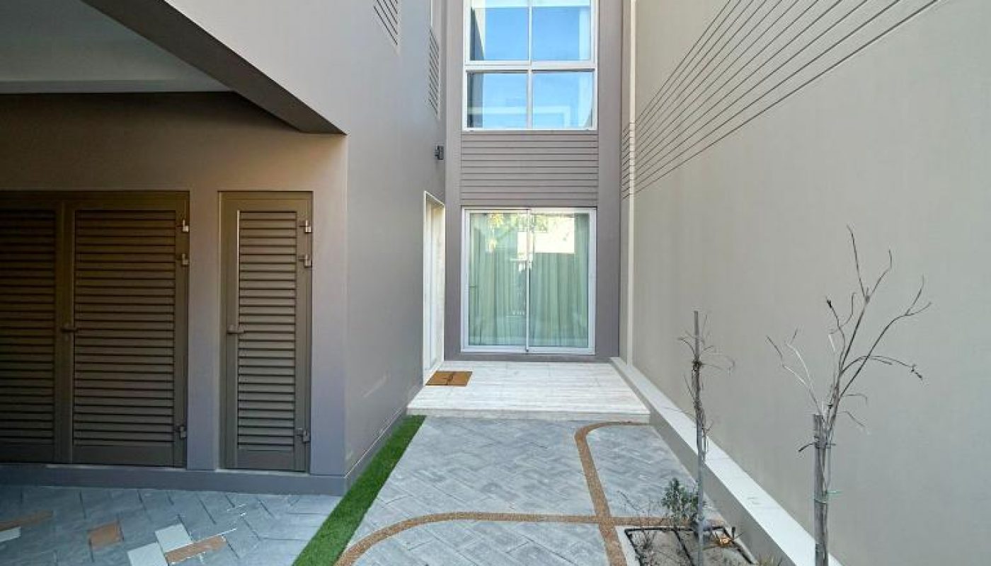 A modern apartment building exterior featuring a paved walkway, planted strip with young trees, and a glass door entryway.