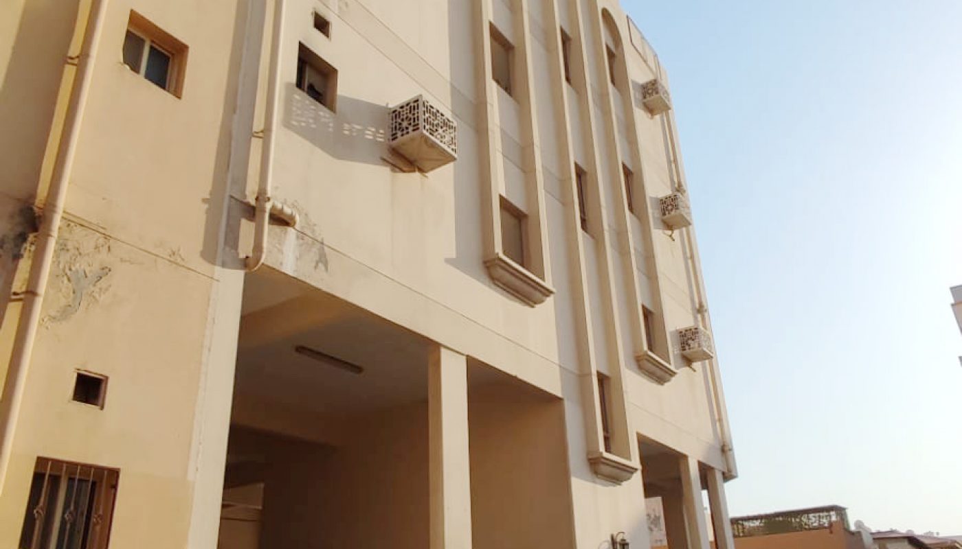 Four-story beige and white residential building with balconies, parking area on the ground floor, and a car parked underneath, set on a narrow street under a clear sky.