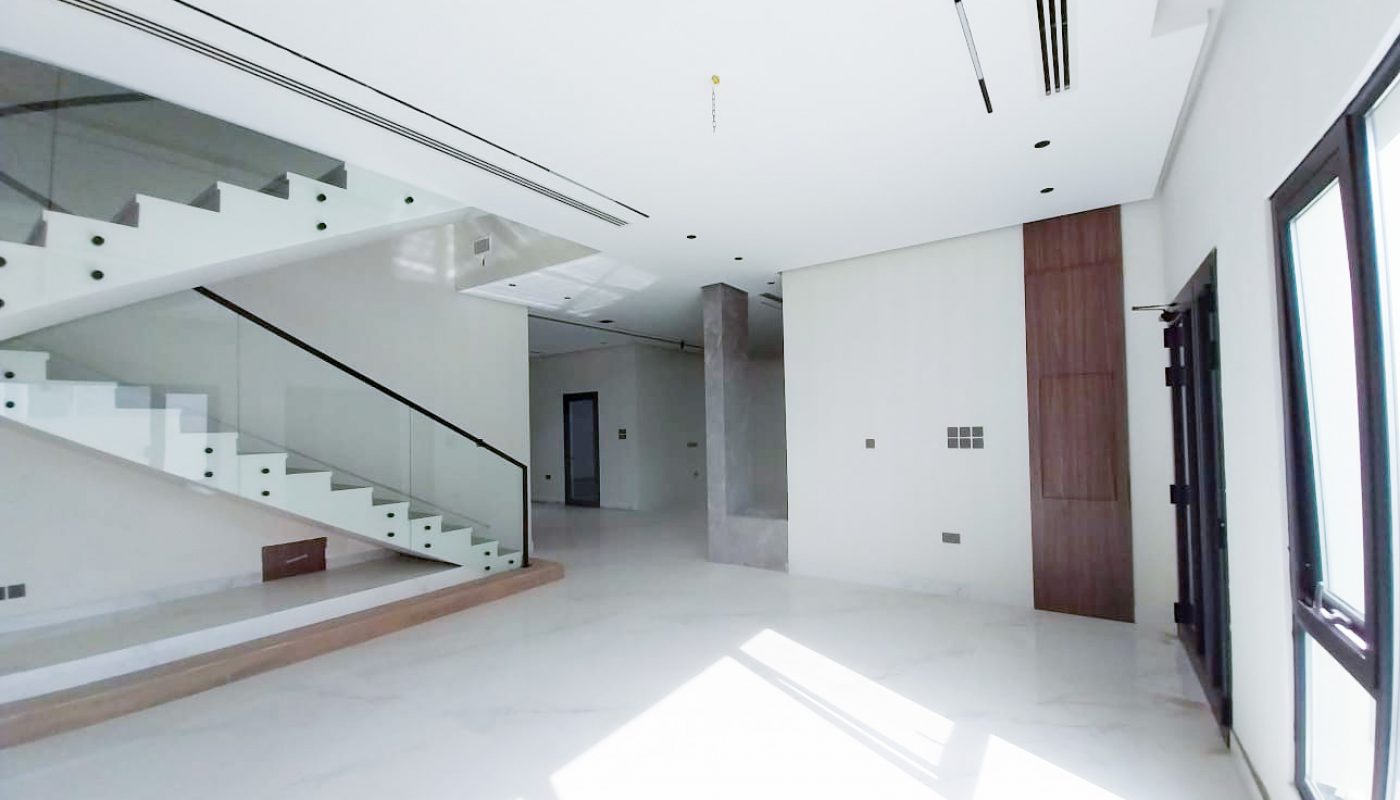 Modern, unfurnished interior with white marble floors, large windows, glass stair railings, and minimalistic design elements. Sunlight streams through the windows onto the floor.