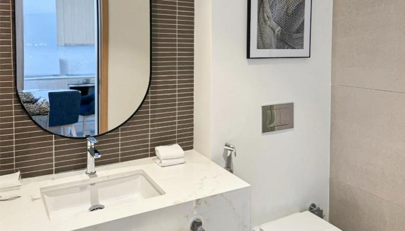 Modern bathroom with a large mirror above a sink, a wall-mounted toilet, framed artwork, and towels neatly arranged on a shelf below the countertop.