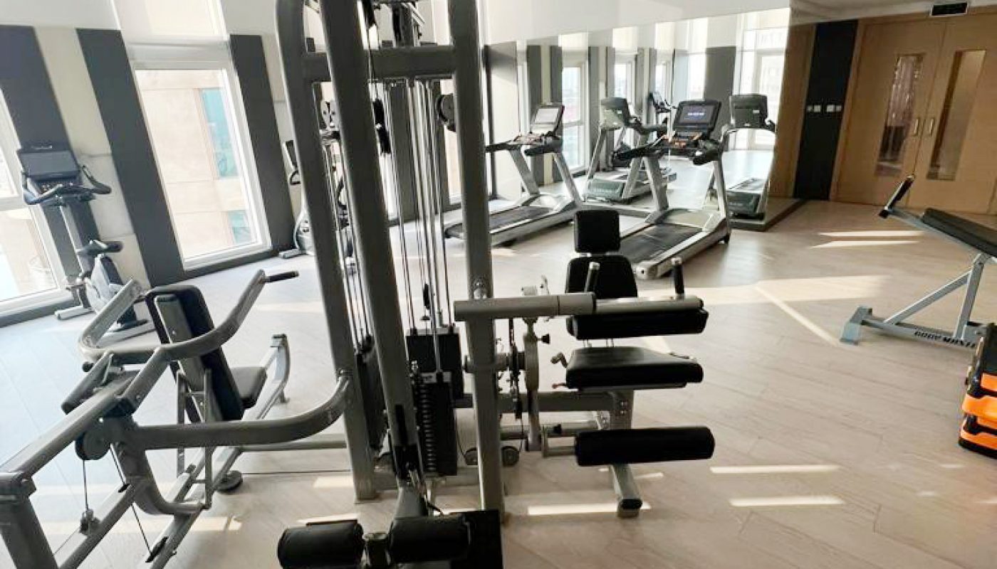 A well-lit gym with various exercise machines, including treadmills, weight benches, and resistance equipment, on light-colored wood flooring.