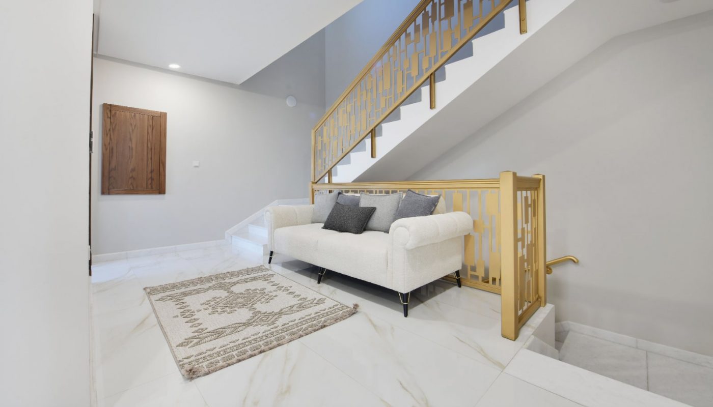 Modern interior with white marble flooring, a white sofa, patterned rug, and staircase featuring gold-toned railings. Walls are light-colored with minimal decor.
