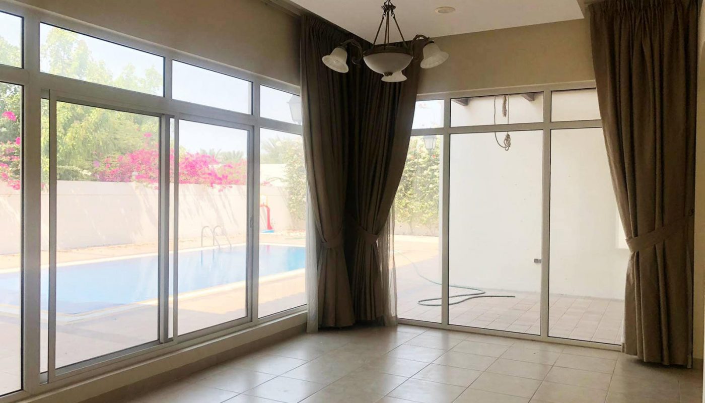 A bright, empty room with large windows and curtains, a hanging light fixture, and a view of an outdoor pool and garden through the glass doors in this spacious villa for rent in Hamala compound.