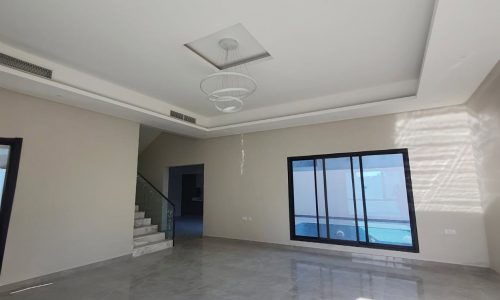 A spacious, empty living room with large tile flooring, off-white walls, a central modern chandelier, a staircase on the left, and large windows allowing natural light to stream in like an auto draft.