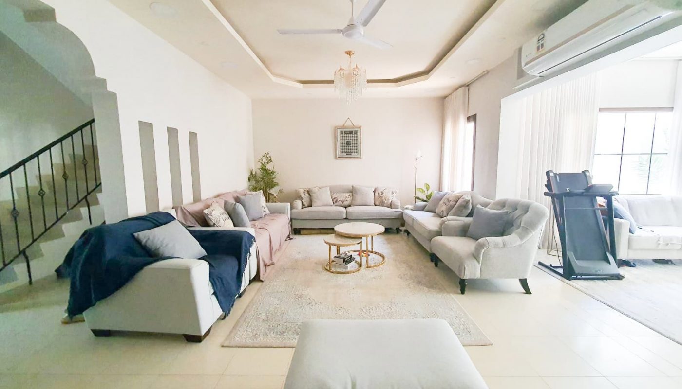 A spacious living room with a U-shaped sectional sofa, a treadmill, and a ceiling fan. A chandelier hangs from the ceiling, and there's a large window with sheer curtains on the right.