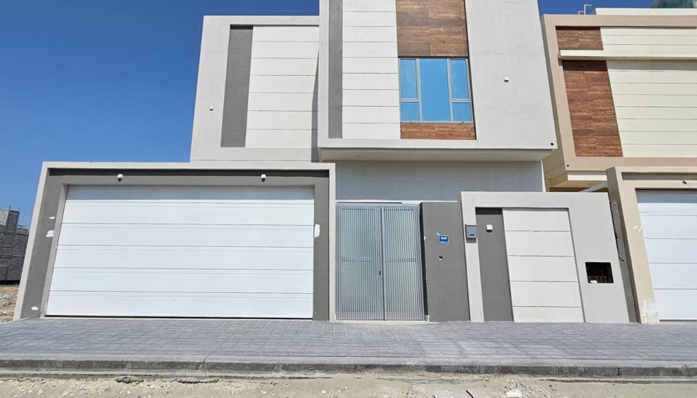 Modern two-story house with a minimalist design, featuring white and beige panels, large windows, and a double garage door. Located on a newly paved street with a clear blue sky above.