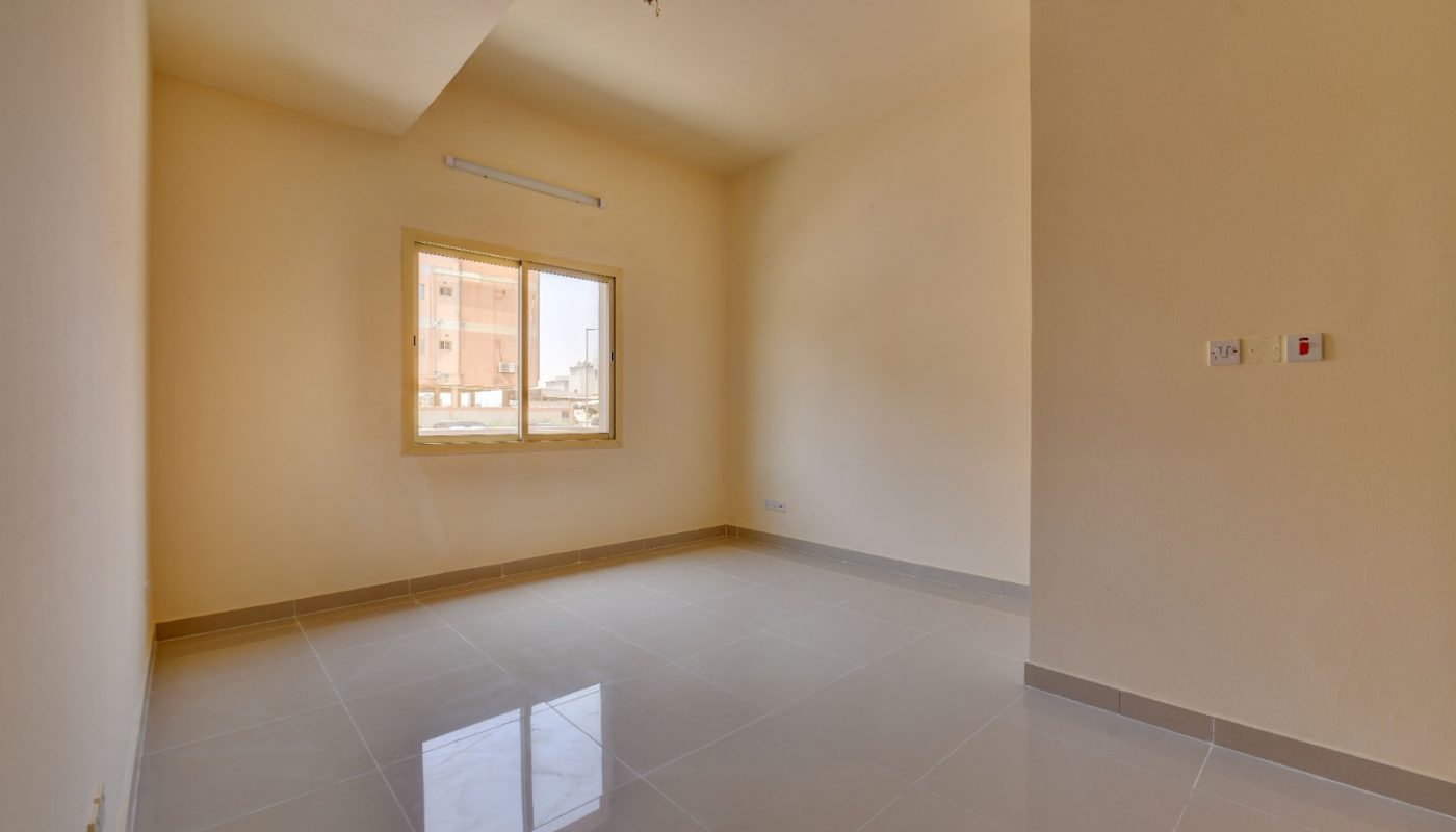 Empty room with cream-colored walls, tiled floor, a single window letting in natural light, and a few electrical switches on the right wall.