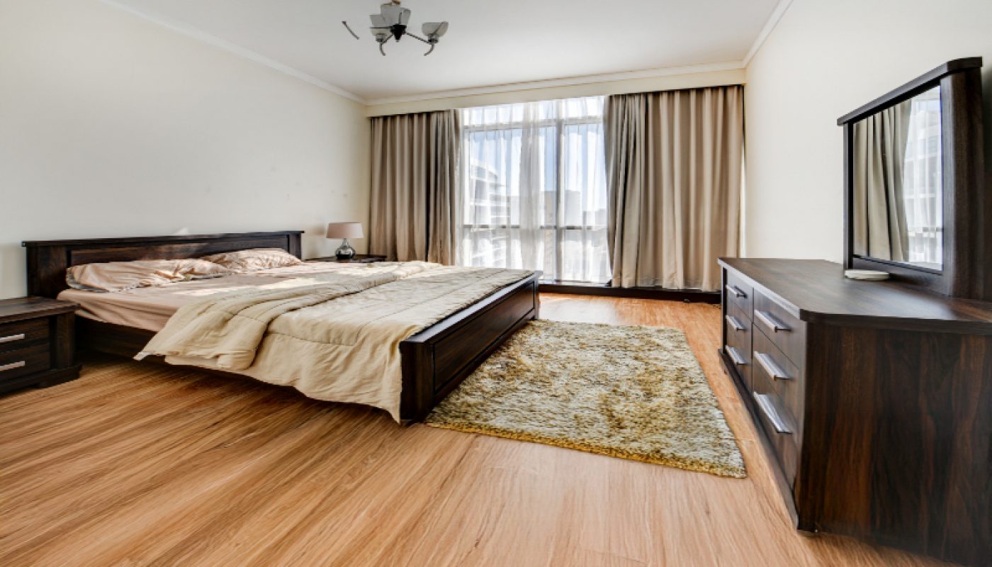 A modern bedroom with a wooden bed, beige bedding, a nightstand, dresser with mirror, large window with curtains, and hardwood floors.
