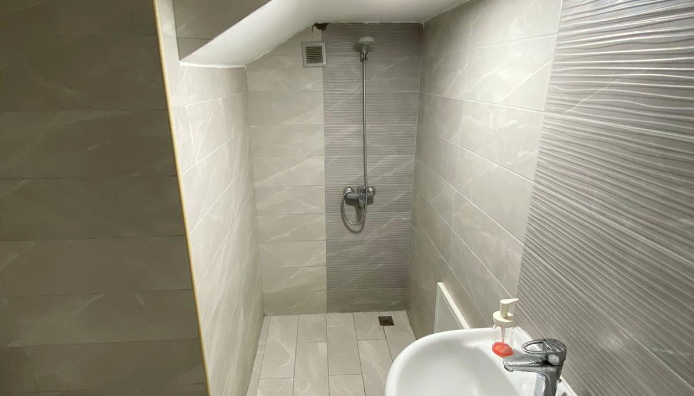 A small, tiled bathroom with a wall-mounted sink, a faucet, a toilet in the corner, and a shower area under a sloped ceiling.